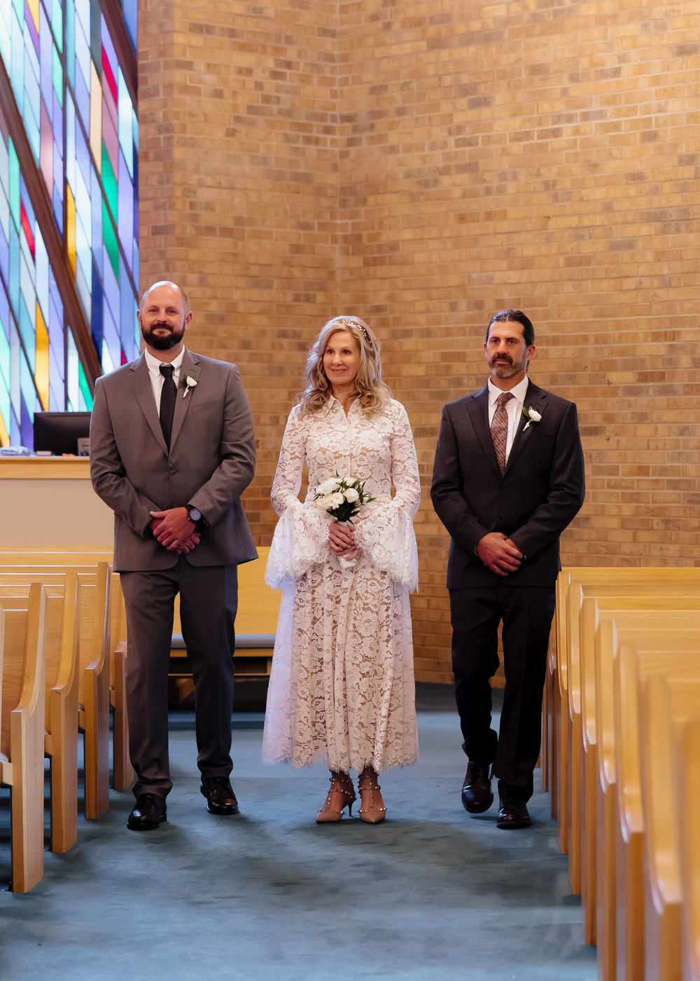 Bride in white lace midi dress walks down church aisle flanked by two groomsmen during wedding ceremony.