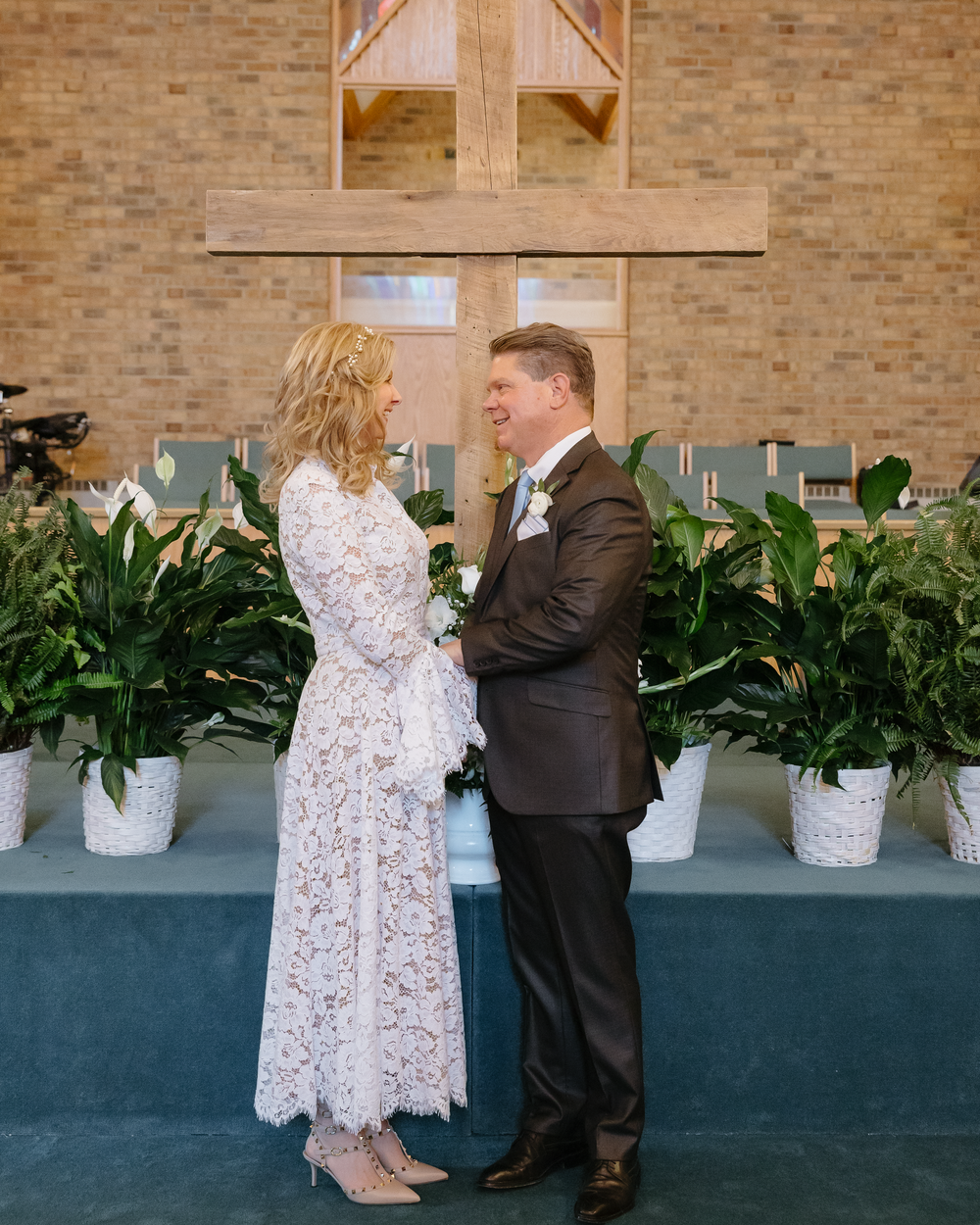 Bride and groom exchange vows at church altar decorated with white potted plants beneath wooden cross.