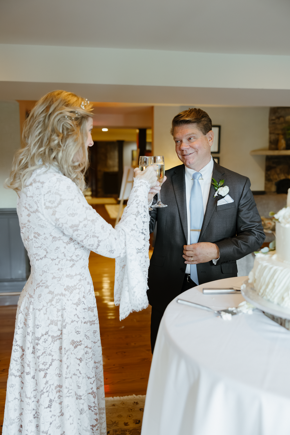 Bride and groom toast with champagne glasses beside white wedding cake at reception celebration.