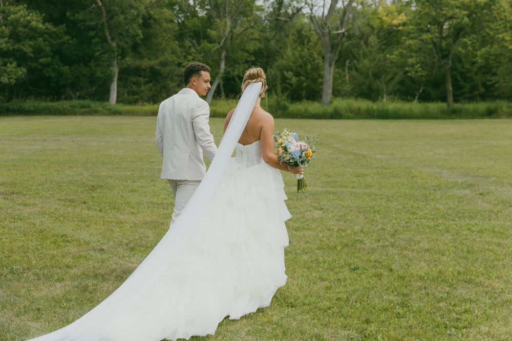 A couple in wedding attire walks together on a lush green lawn surrounded by trees.