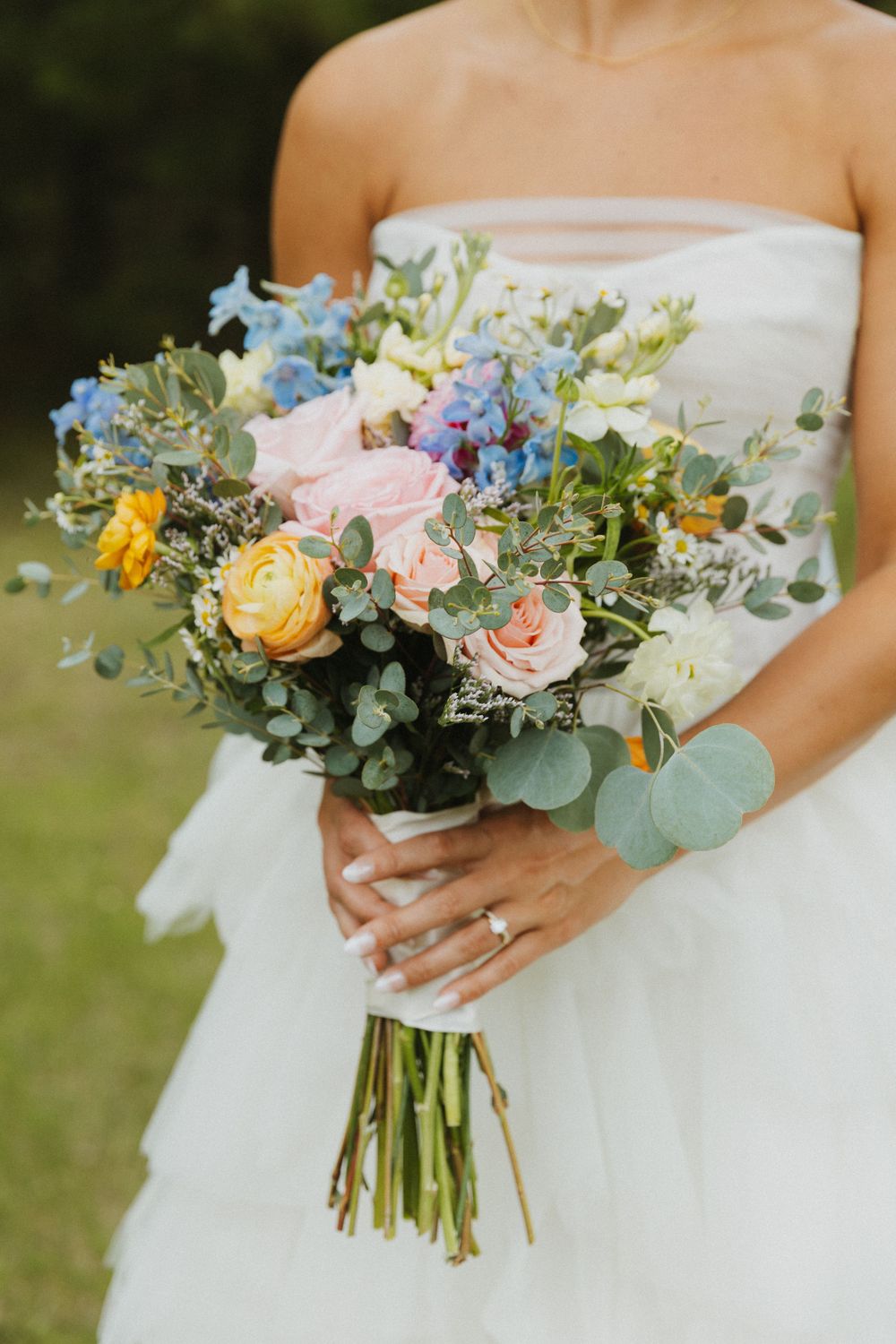 Beautiful wedding bouquet with pastel roses, eucalyptus, and blue flowers against a white dress.