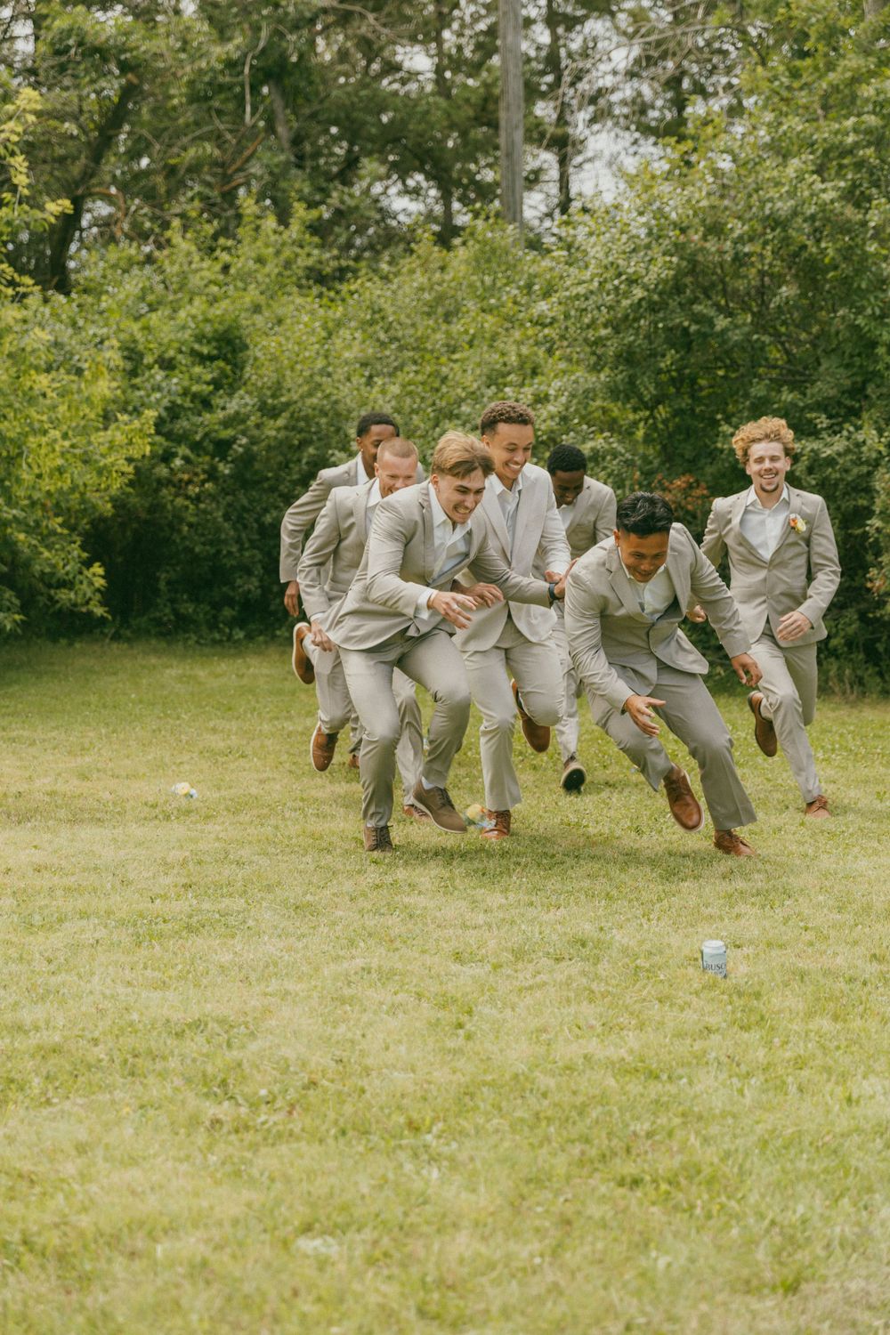 Groomsmen in light gray suits playfully run across a green lawn during wedding festivities.