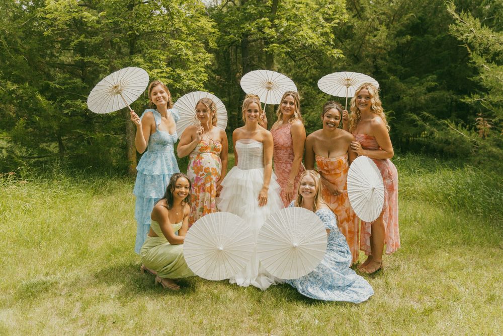 Wedding party with parasols poses together on grass in pastel dresses.