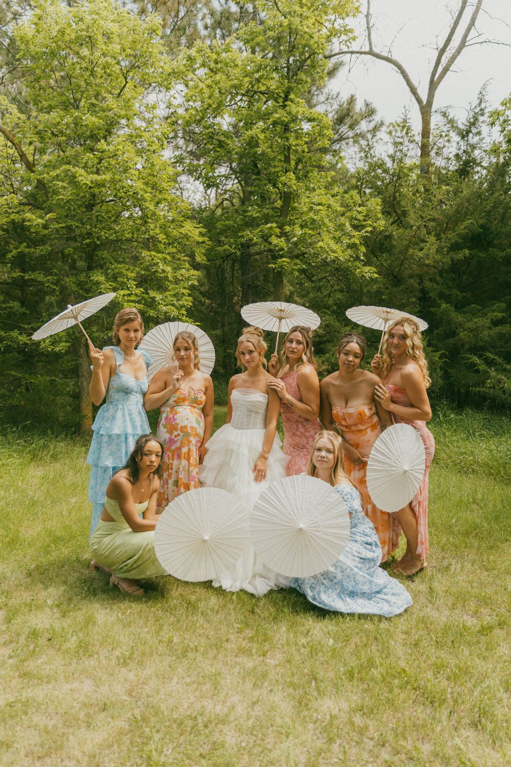 Bridesmaids in floral dresses pose with white parasols on a sunny lawn.