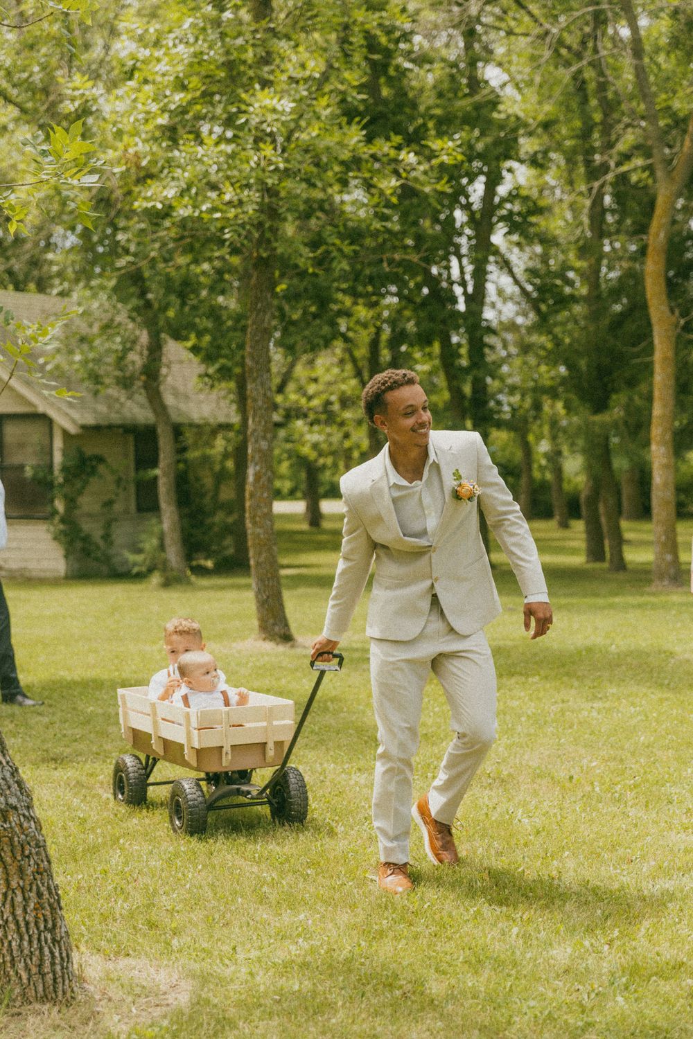 A person in a white suit pulls a rustic wooden wagon across a sunny lawn with trees.