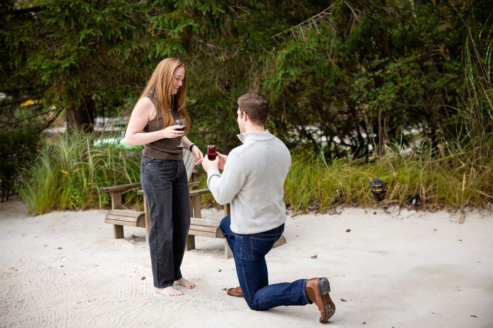Man kneeling on sandy beach proposing to woman with ring box outdoors.