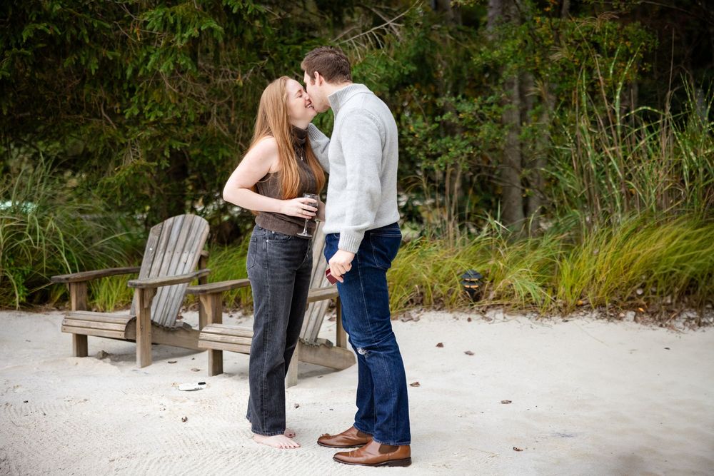 A couple shares a romantic kiss while holding hands on a sandy path surrounded by lush greenery.