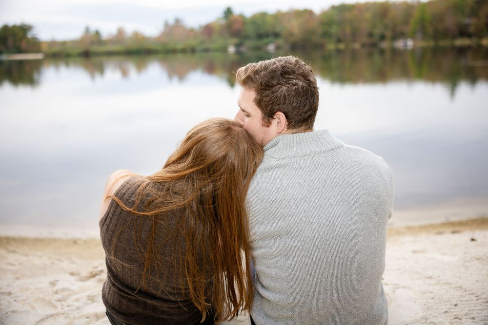 A romantic couple sits together facing a serene lake, the woman resting her head on the man's shoulder.