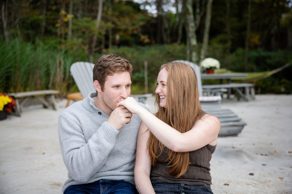 A smiling couple sits on a sandy beach, the man kissing the woman's hand during an engagement photo session.