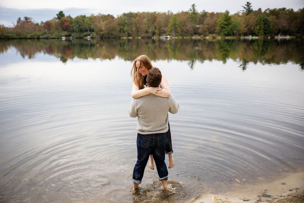 Couple embracing from behind while standing in calm lake with autumn trees reflected.