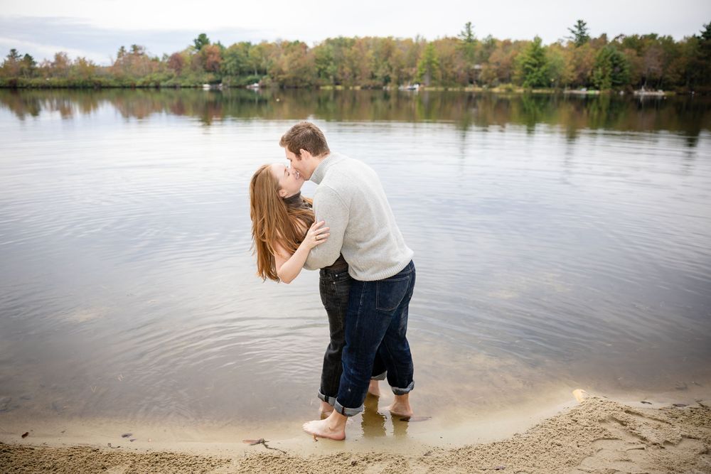 Couple kissing while standing in shallow lake water with autumn trees in background.