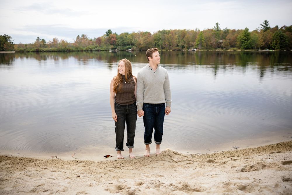 Couple holding hands barefoot on sandy lake shore with autumn trees and cloudy sky.
