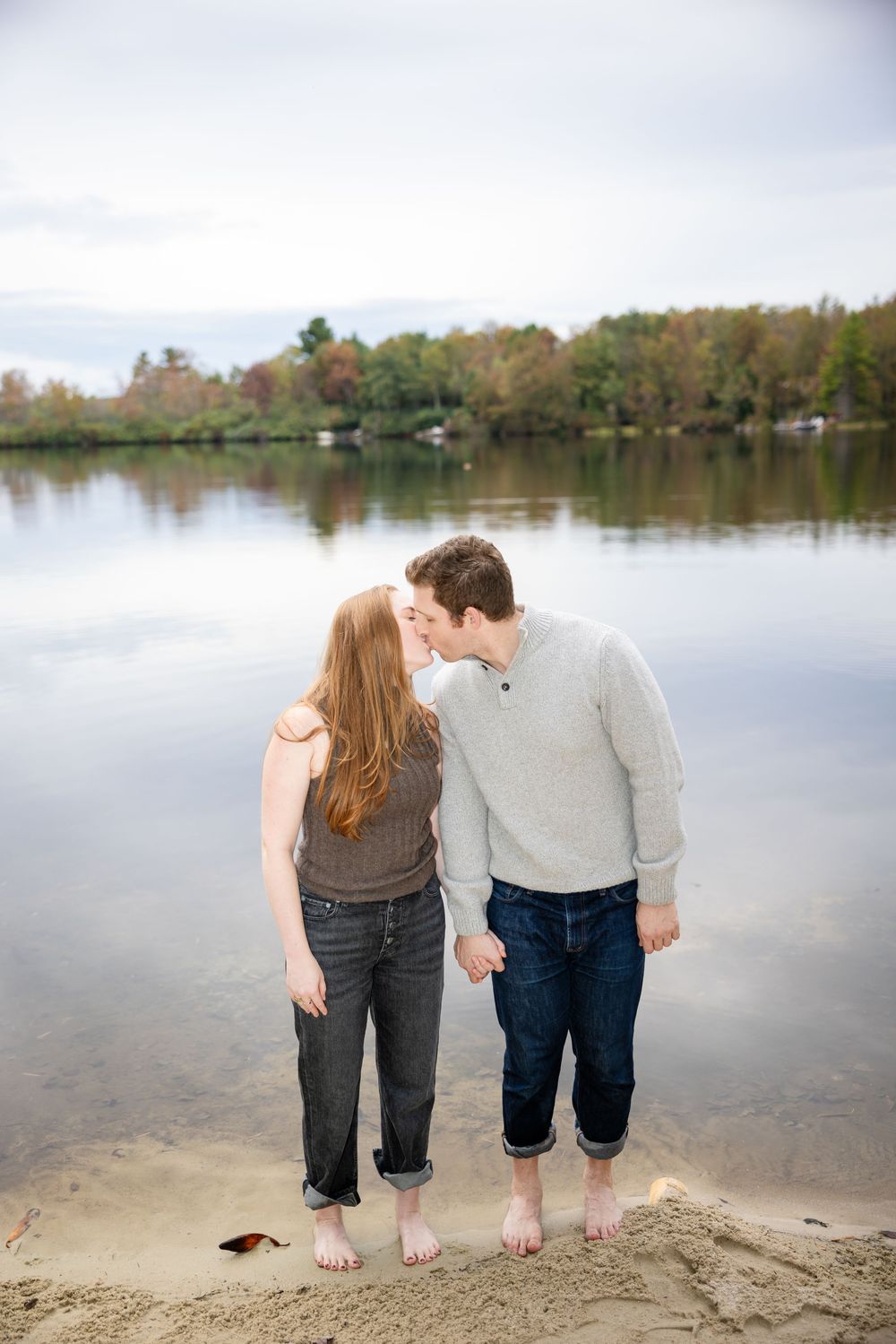 Couple kissing barefoot on sandy lake shore with colorful autumn trees reflected in water.