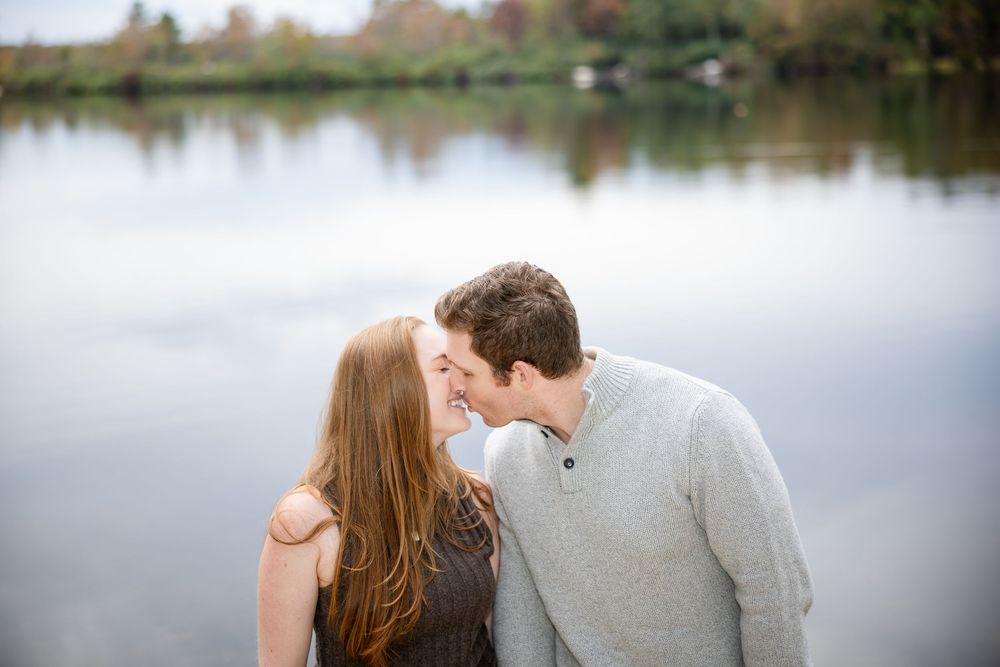 Couple sharing a romantic kiss near a calm lake surrounded by autumn foliage.