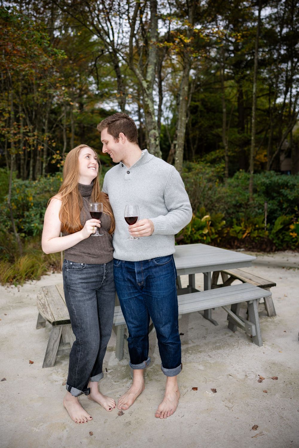 Couple barefoot on sandy beach holding red wine glasses smiling at each other with picnic table and autumn trees behind.