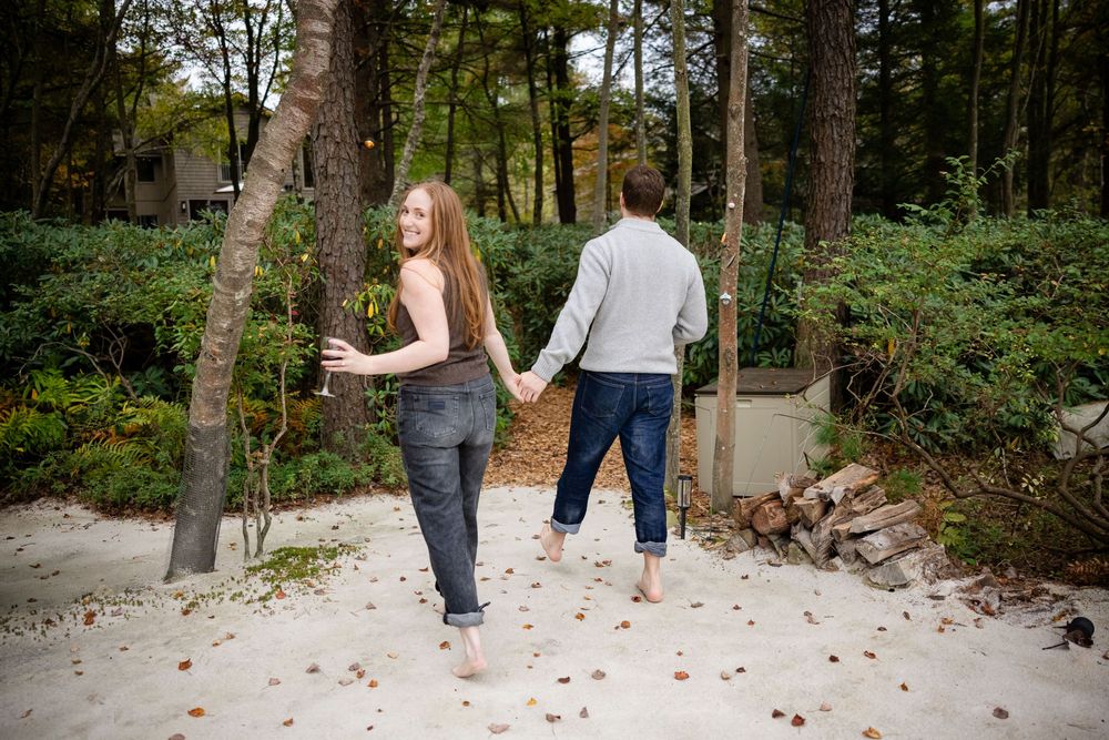 Couple holding hands walking barefoot on sandy path through wooded area.