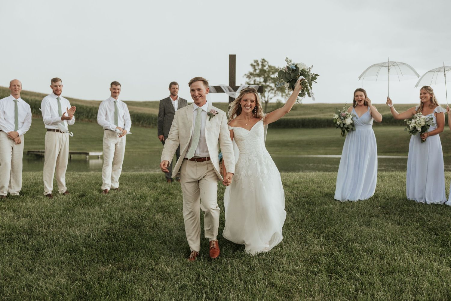 A wedding party stands on a green lawn with bridesmaids in light blue dresses and groomsmen in beige suits.