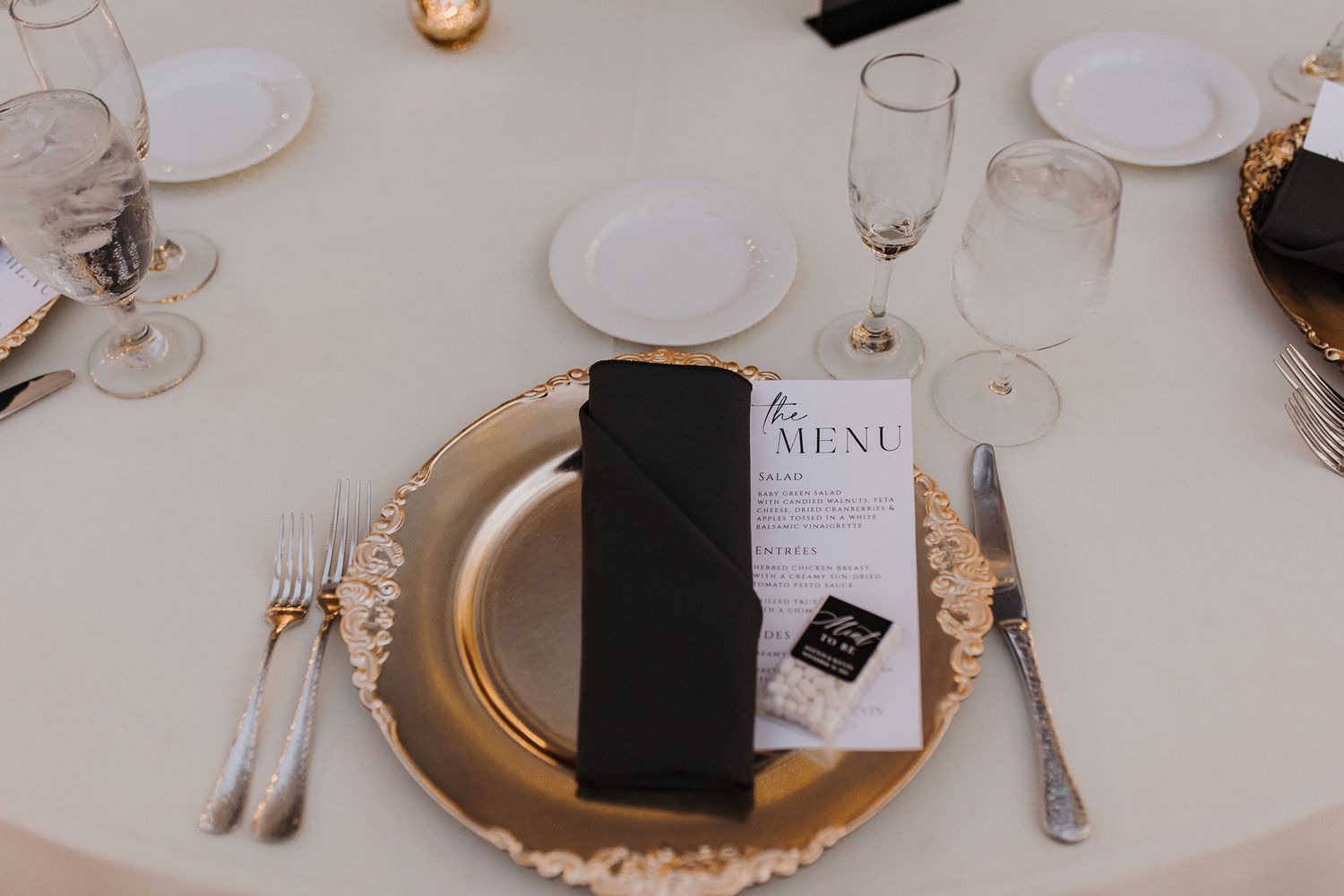Elegant place setting with gold charger plate, black napkin, menu card, and formal silverware on white tablecloth at Lake Oak Meadows.