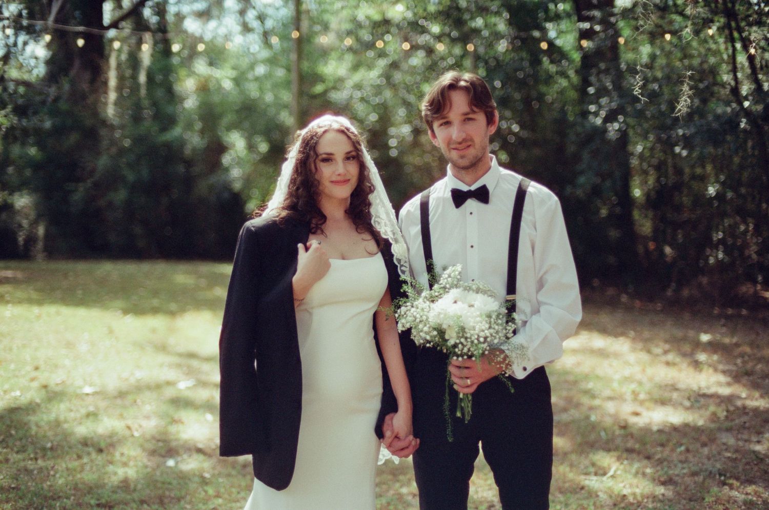 A couple in wedding attire poses together in a sunlit forest setting during their outdoor wedding celebration.