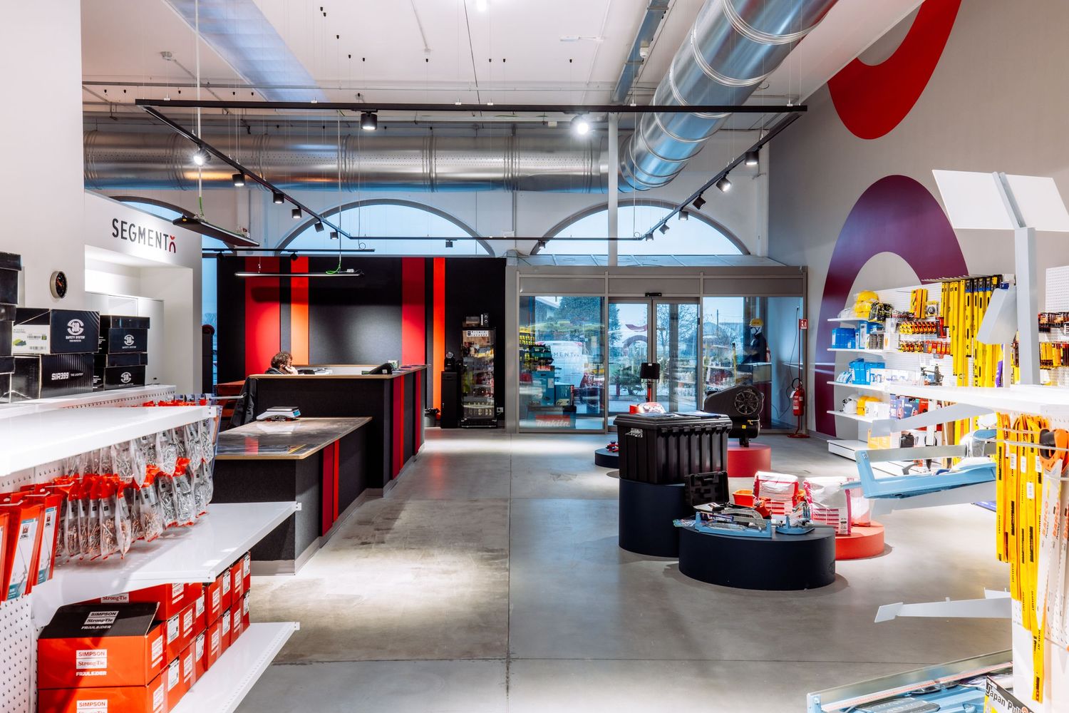 Modern retail store interior with red accents, industrial ceiling, and sleek merchandise displays along concrete floors.