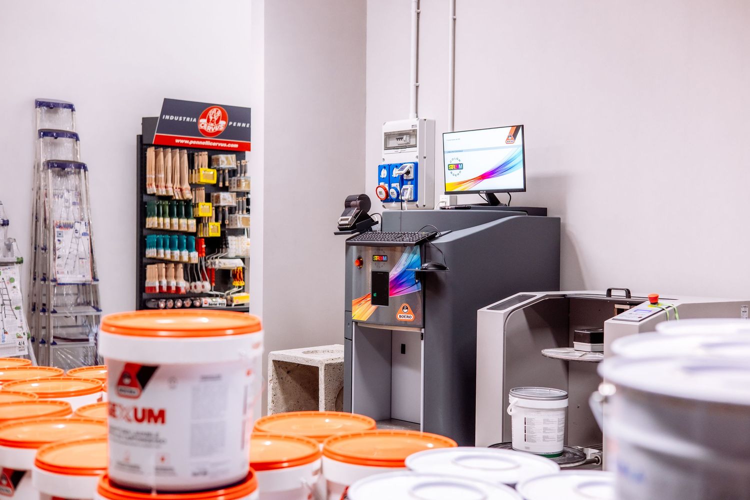 Interior view of a print shop workspace with computer setup, orange buckets, and organized supplies on shelves.
