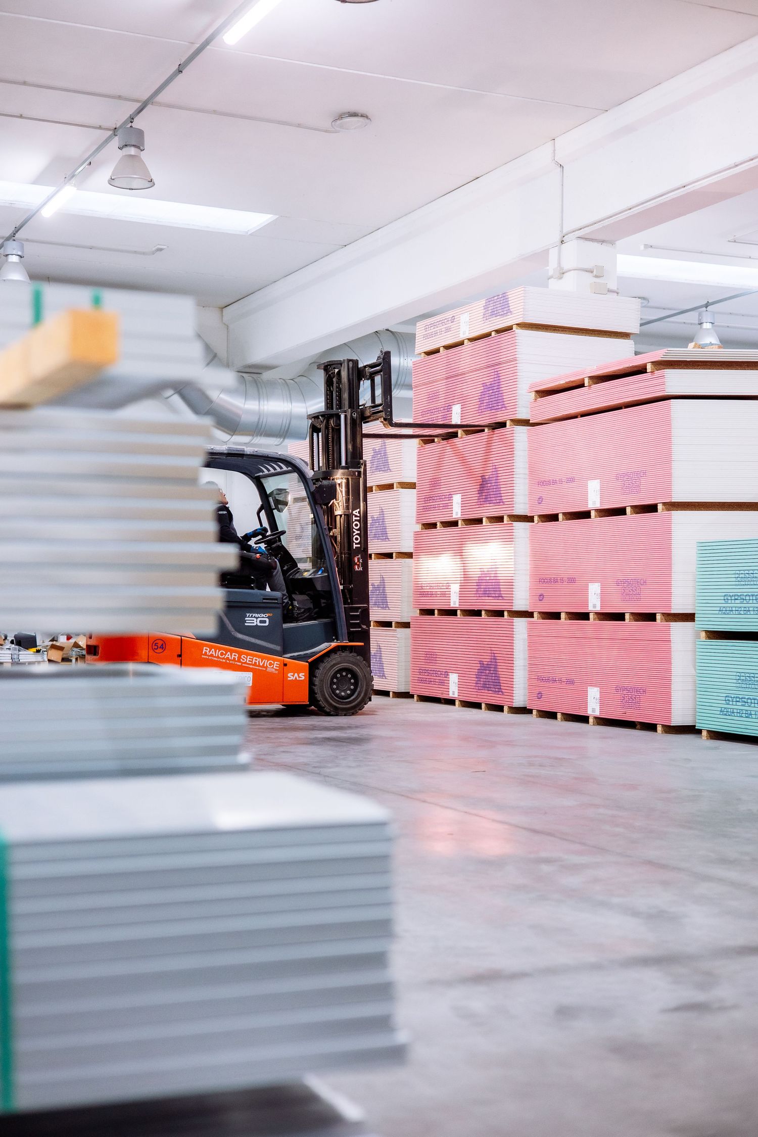 Orange forklift operating in warehouse with stacked pink storage containers and gray materials on concrete floor.