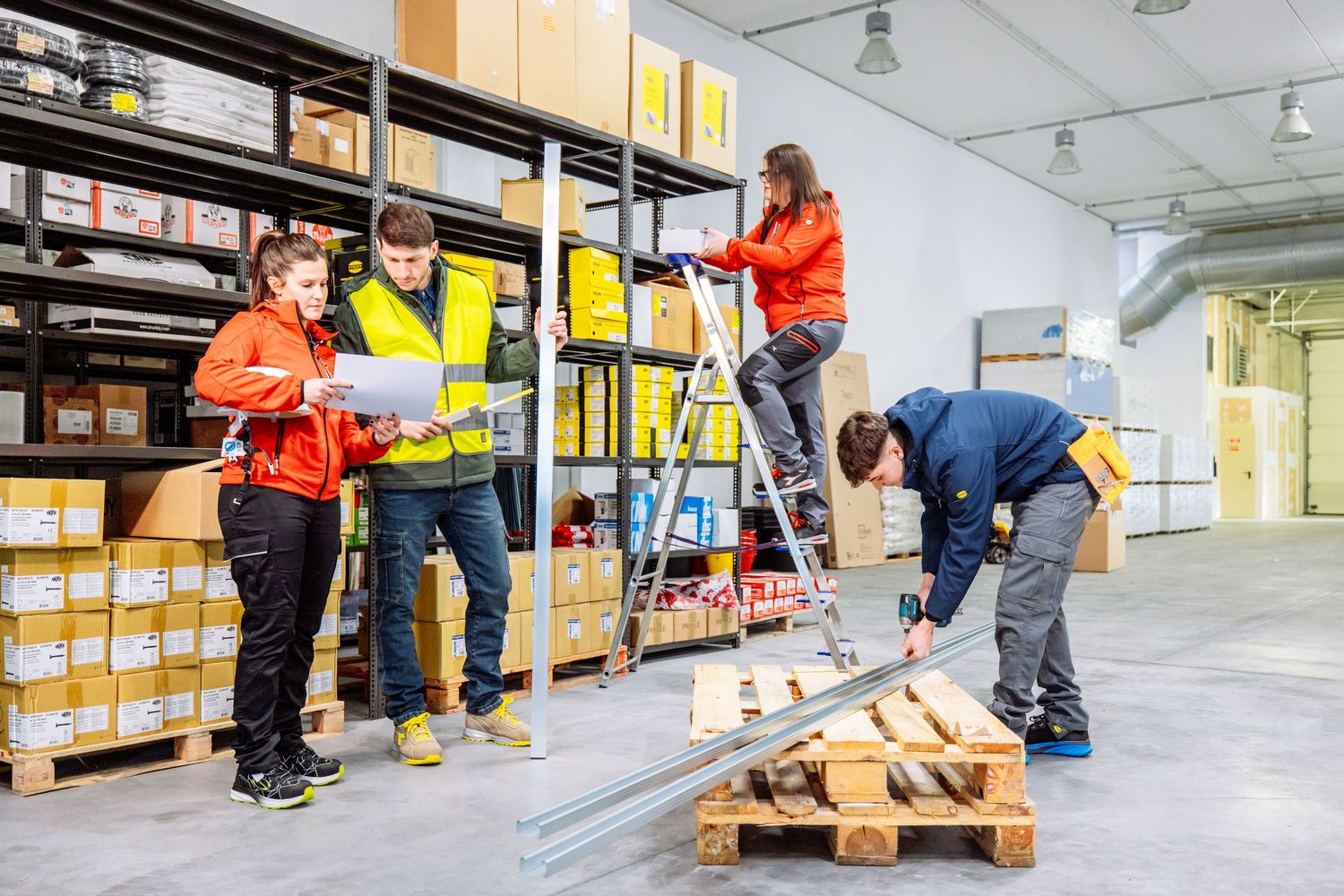 Workers in warehouse organize and move boxes on wooden pallets using pallet jacks and ladders.