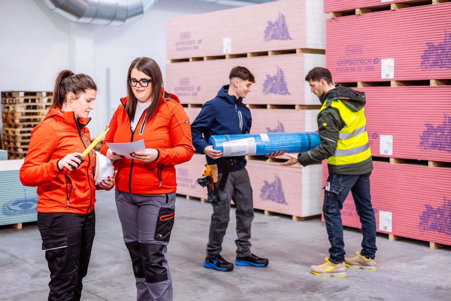 People in red jackets work together to assemble and stack pink insulation panels in a warehouse setting.