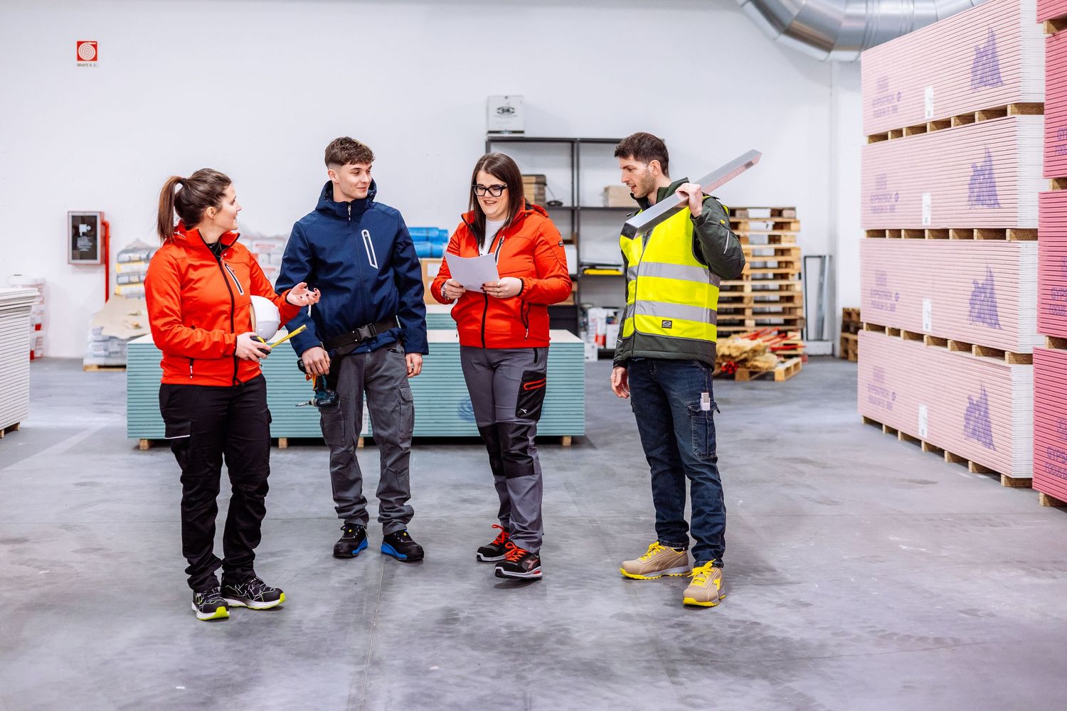 A group of workers in safety gear stand together in a warehouse facility with pink walls and pallets.