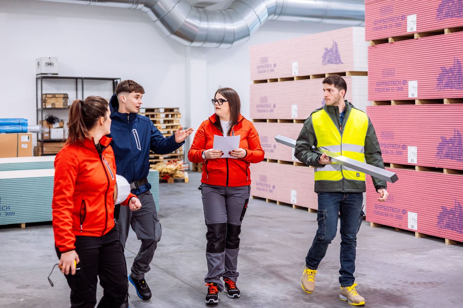 Workers in red jackets and safety vests inspect packages in a warehouse facility with pink walls.