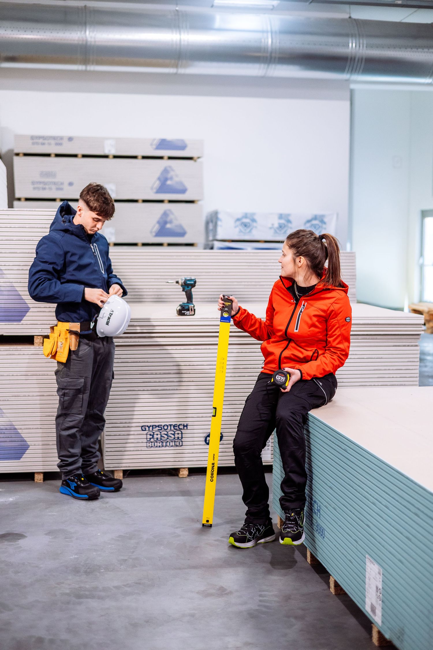 Two workers in winter jackets taking measurements and inspecting building materials in a warehouse setting.