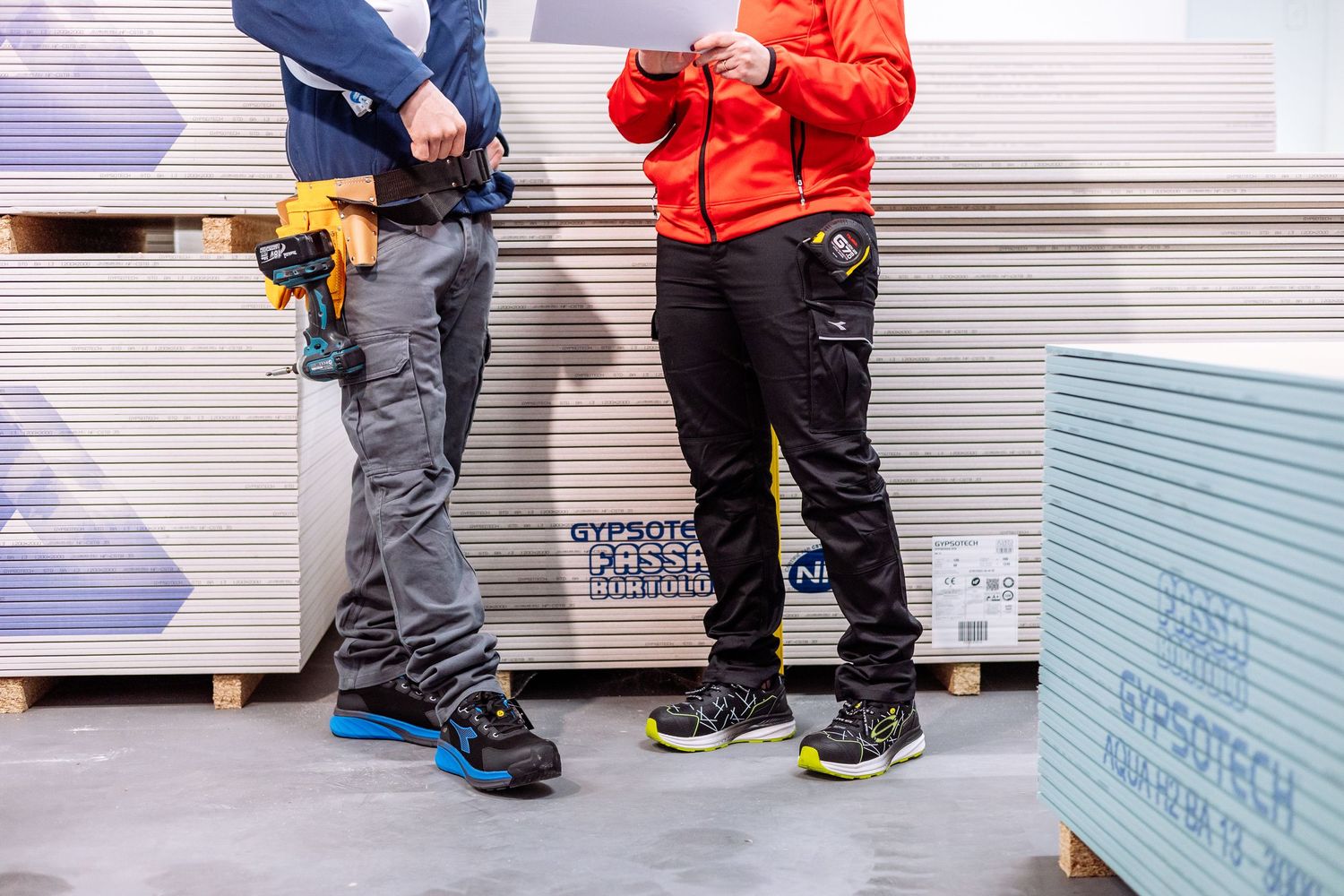 Two workers in protective workwear and safety boots stand next to stacked construction materials in a warehouse.