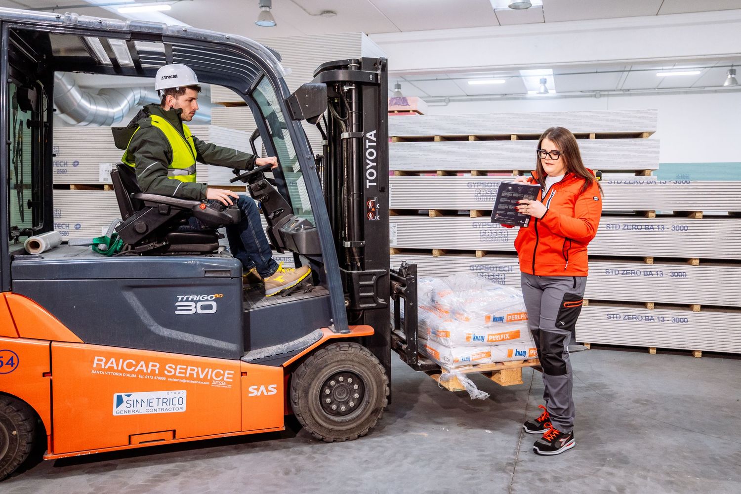 Orange forklift trucks operate in a bright warehouse facility with stacked pallets and workers in safety gear.