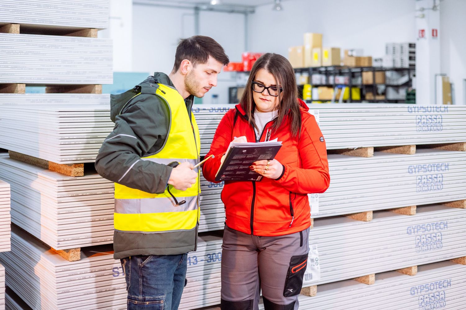 Two warehouse workers review inventory information on a tablet while standing among stacked pallets in a storage facility.
