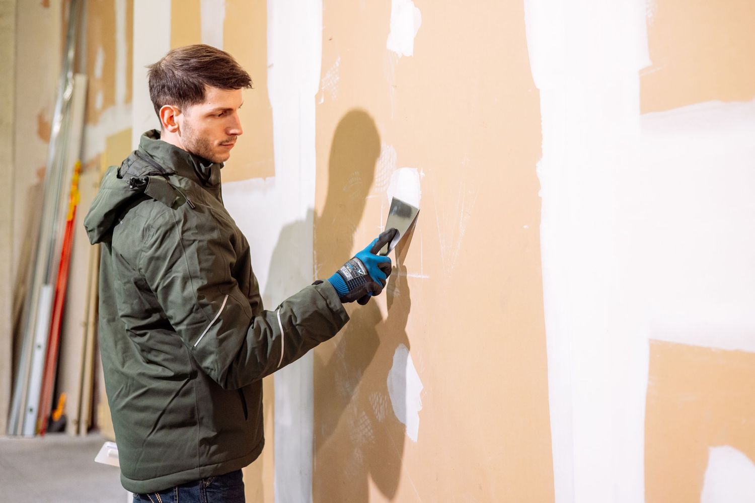 A person in a dark winter jacket and gloves uses a scraper tool on a white wall during renovation work.