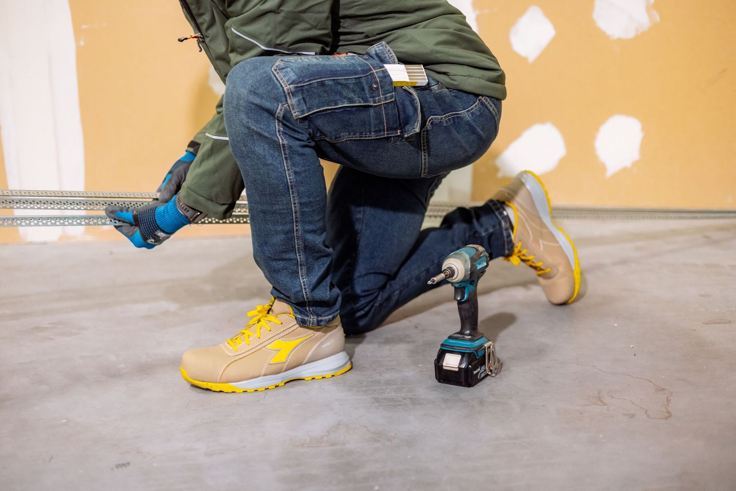 Person wearing jeans and sneakers kneels on concrete floor while performing construction or installation work.