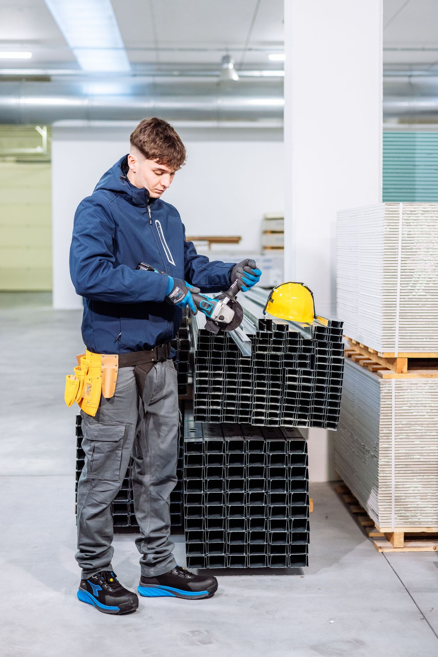 Worker in blue jacket and gray pants inspecting metal construction materials in a warehouse setting.