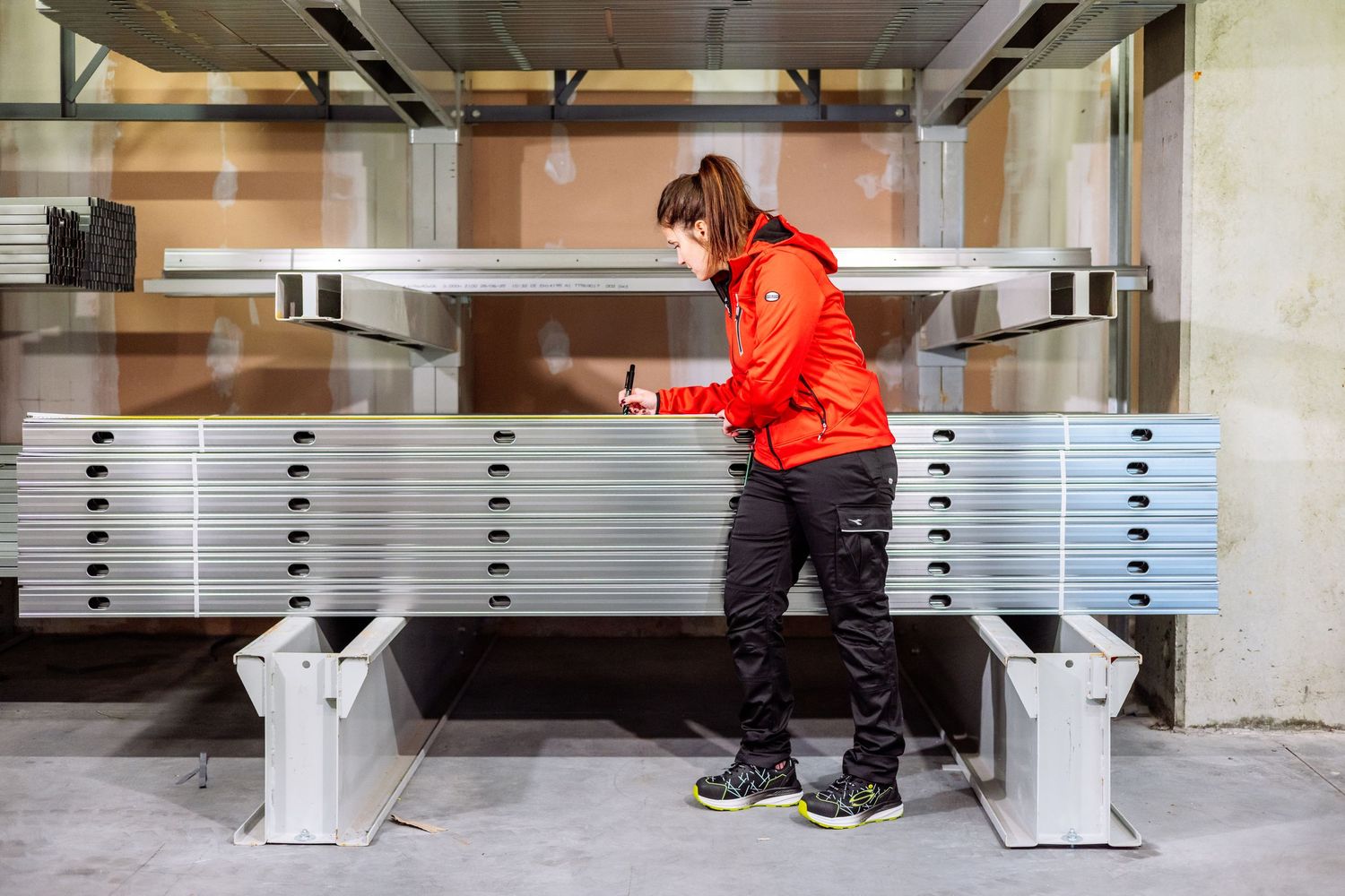 Worker in bright orange jacket organizes stacked aluminum construction materials in industrial storage area.