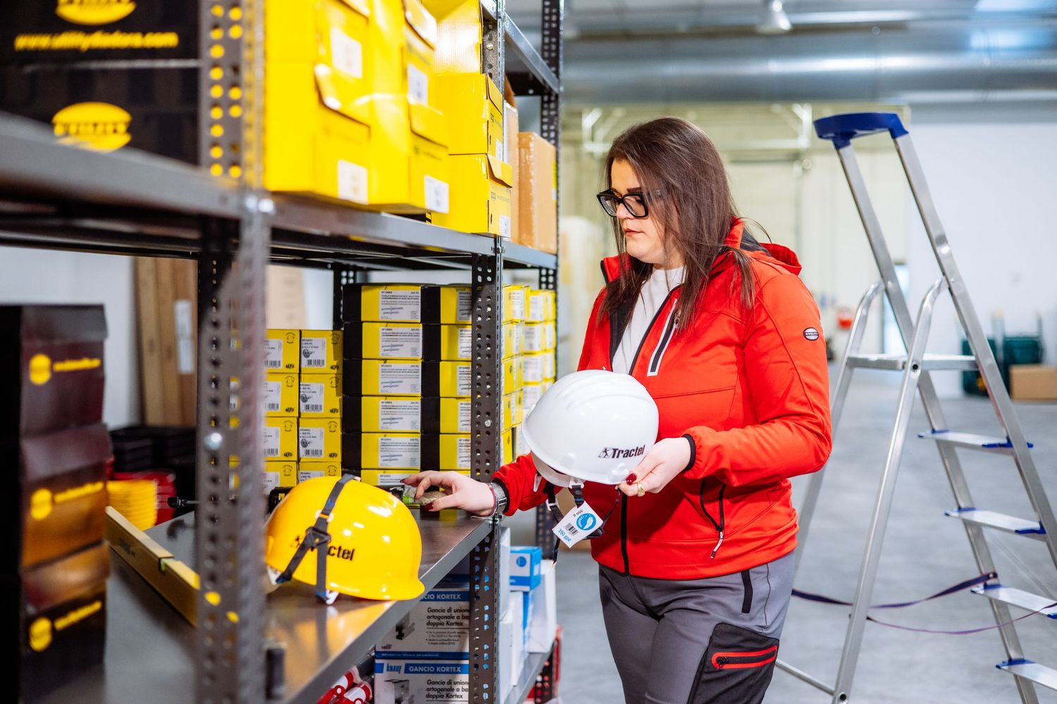 Worker in red winter jacket inspects safety equipment on industrial storage shelves with yellow bins and ladder nearby.