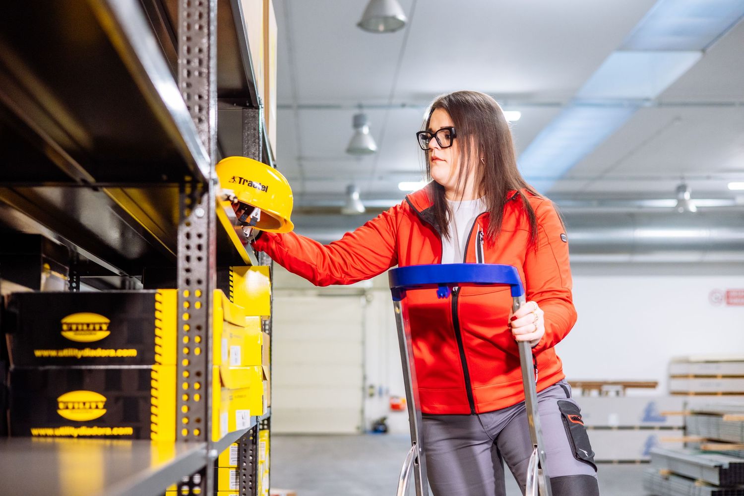 Warehouse worker in red jacket scanning inventory items on industrial shelving with handheld device.