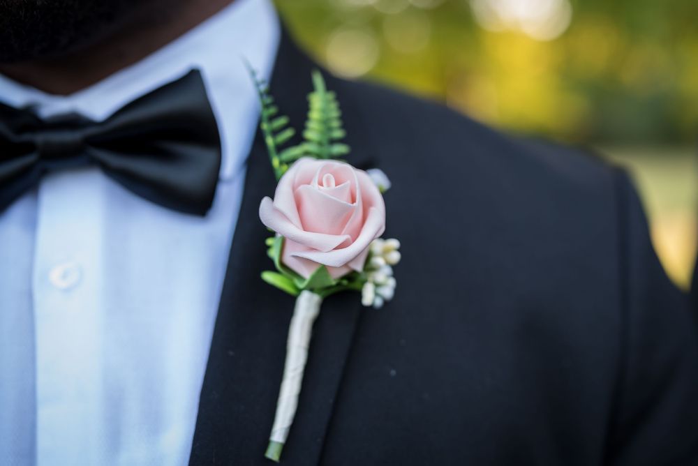A pink rose boutonniere with delicate fern leaves pinned to a black tuxedo lapel in soft natural lighting.