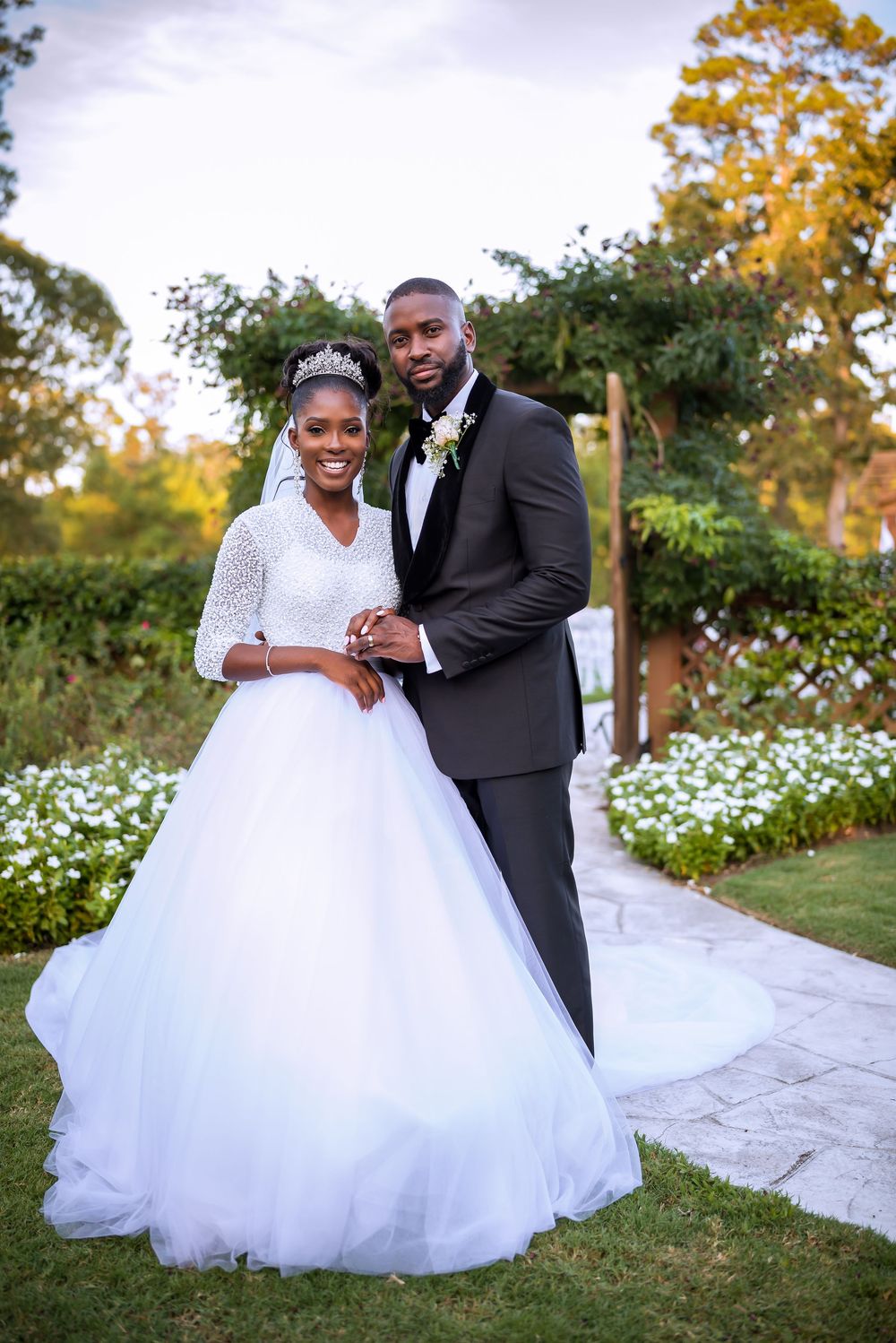 A beautiful wedding portrait outdoors with a white ballgown dress and black tuxedo against a garden backdrop with white flowers.