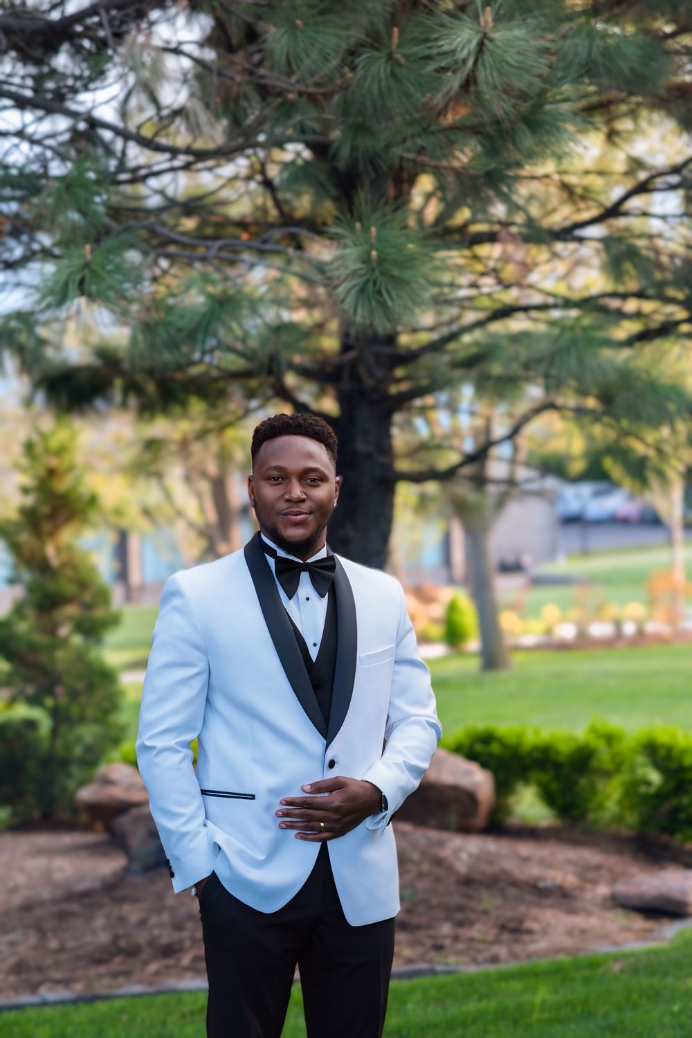 A groom in a stylish white dinner jacket with black lapels and bow tie poses outdoors near evergreen trees.