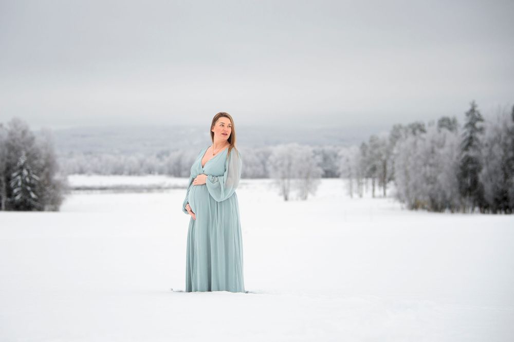 A figure in a flowing mint green dress stands in a serene snowy winter landscape with evergreen trees in the background.