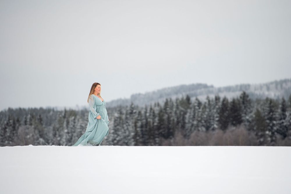 A person wearing a turquoise gown poses elegantly against a snow-covered mountain landscape with pine trees.