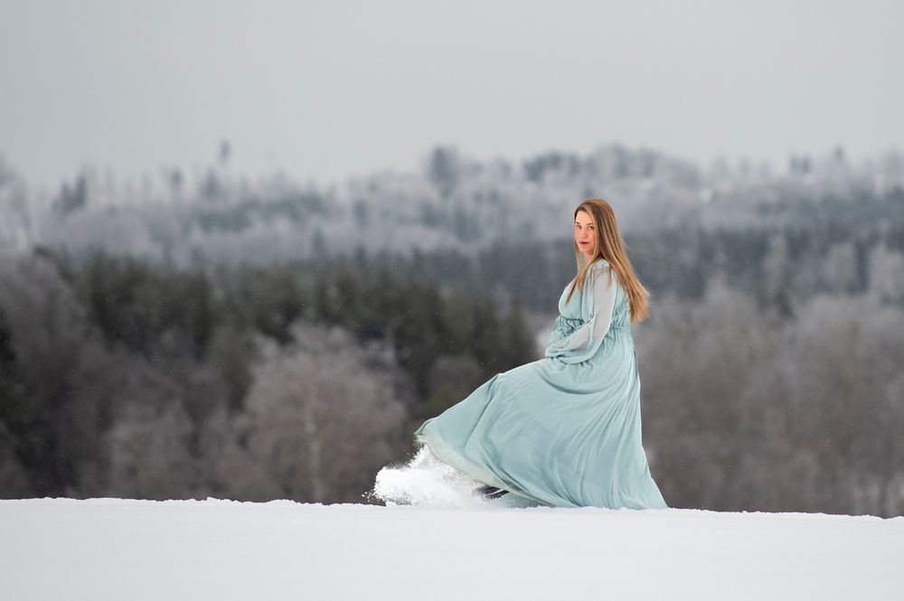 A person in a flowing light blue gown stands gracefully in a snowy winter landscape with frost-covered trees in background.