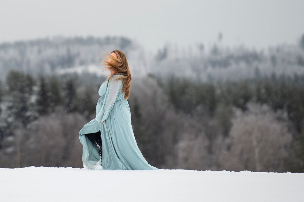 A person in a flowing light blue dress stands in the snow against a backdrop of winter forest and mountains.