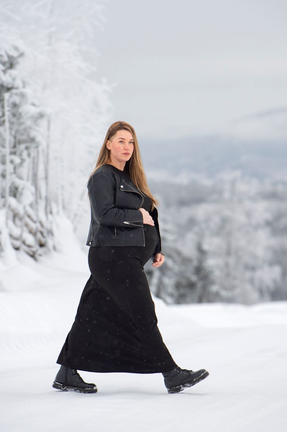 Person in a long black coat poses gracefully against a snowy winter landscape with frost-covered trees.