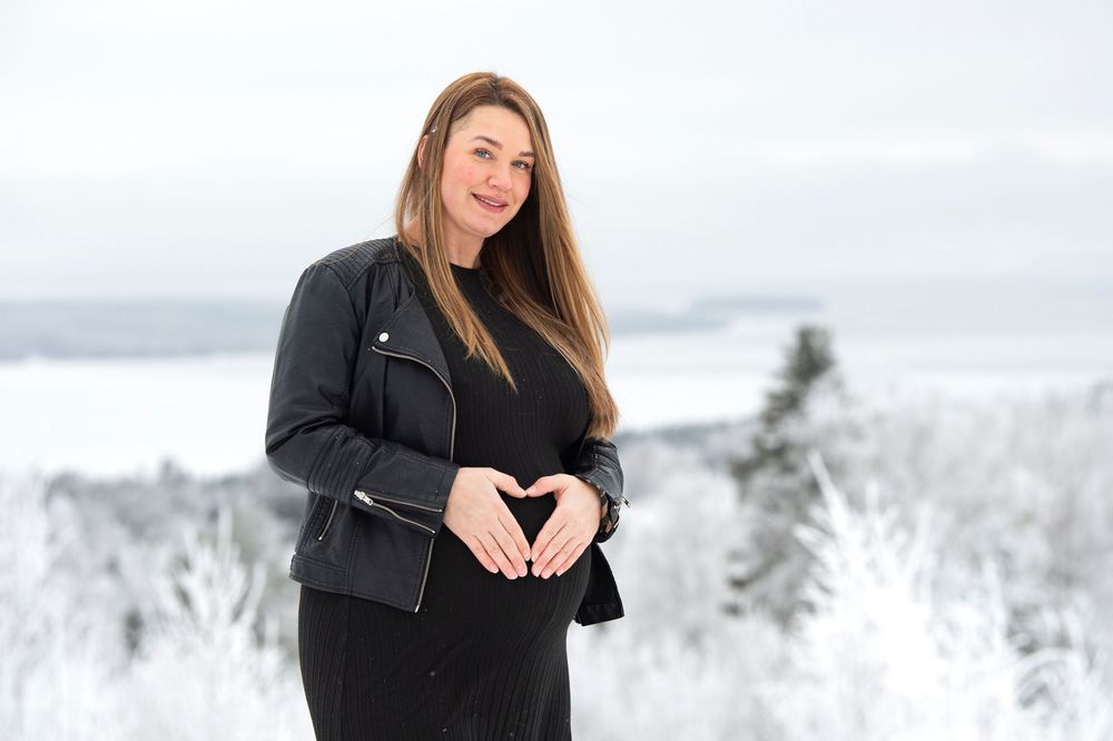 A person in a black jacket poses in a snowy winter landscape with mountains and evergreen trees in the background.