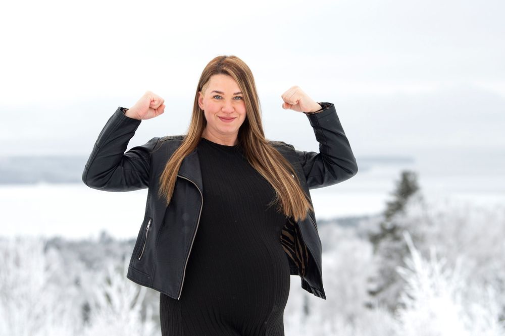 Person wearing a black winter outfit stands in snowy landscape with snow-covered trees in the background.